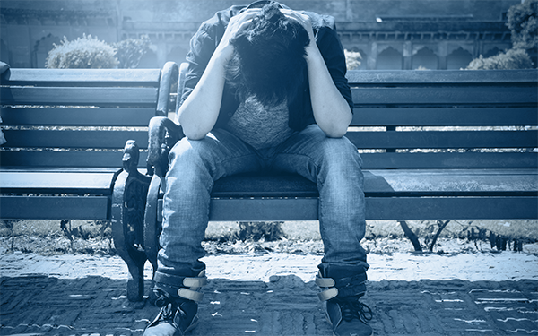 A male student sitting outside on a bench, hunched over with his hands on top of his head.