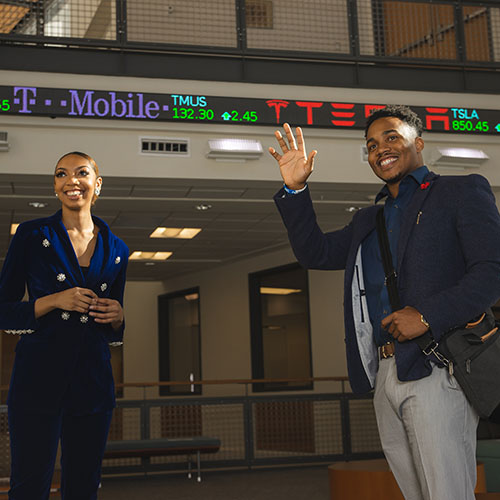 Two students in professional attire standing in front of a slim rolling stock ticker screen.