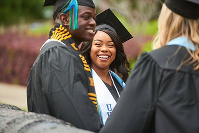 UWF graduates smile while chatting on the Cannon Green as they wear cap and gown.