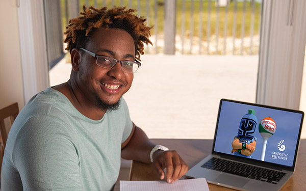 A UWF student smiles and writes with pen and paper by a laptop at home.