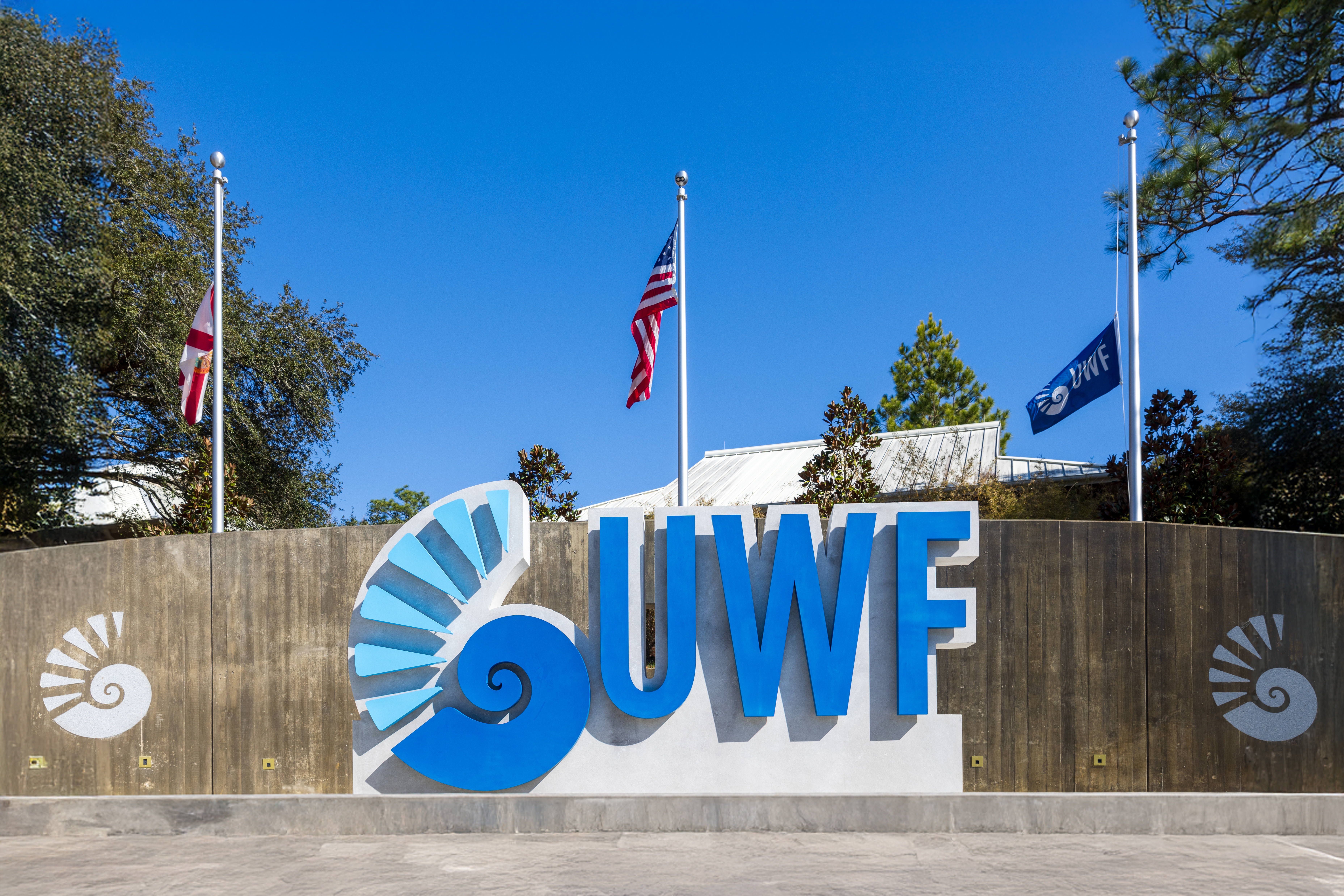 A concrete sign with a nautilus shell and UWF on display in front of a concrete wall and three flag poles.