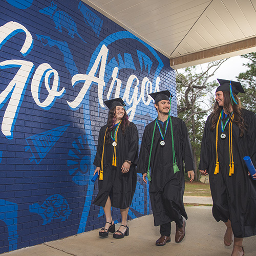 Three UWF grads walking in front of the Go Argos mural