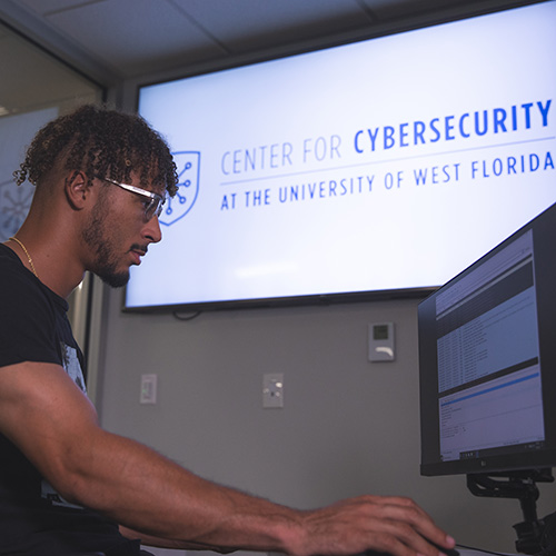 Cybersecurity student using computer with UWF Center for Cybersecurity logo on screen in the background
