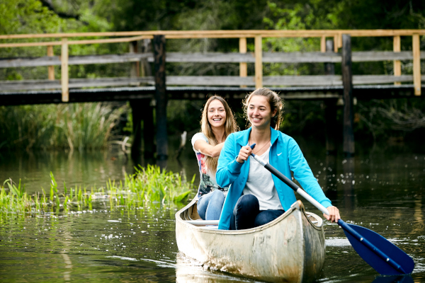 Two UWF students paddling down the river in a canoe with a walkway behind them.