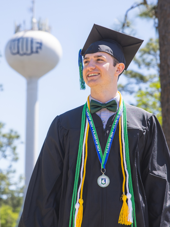 A 2024 graduate in full cap and gown regalia stands in front of the UWF water tower.