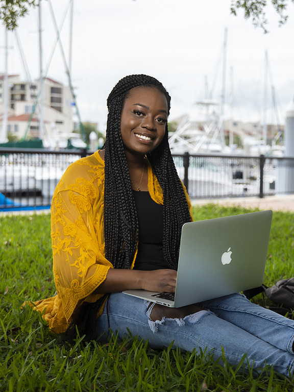 Student sitting on grass using laptop with boats docked in the water in the background.