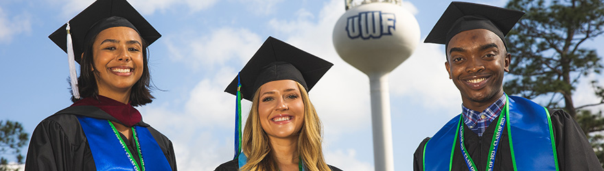 Three graduates smile while wearing cap and gown in front of the UWF water tower.