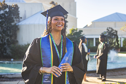 A student standing outdoors in commencement regalia cap and gown attire holding a diploma tube with two people converse in the background while standing in front of a fountain.