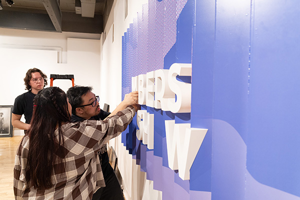 Two students working on a lettered art piece on a wall with a third student standing behind the other two students watching.