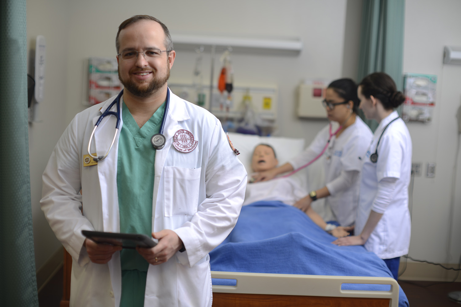 FSU medical student standing in front of a hospital bed where two UWF students are using a stethoscope on a medical dummy model.
