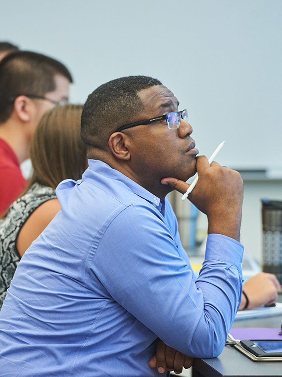 Four students sitting in a row at a table in class with the most forefront student resting their hand on their chin with a pen in their hand.