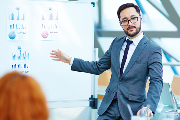 Business professional giving data presentation in front of a board displaying print outs of charts and graphs.